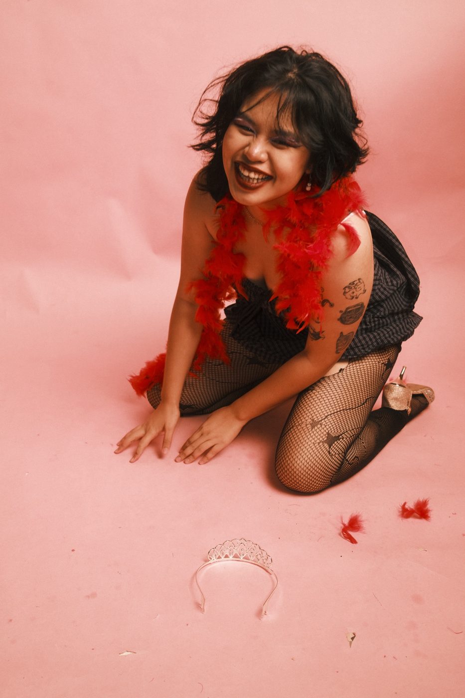 Kneeling and laughing in a red boa, a tiara dropped on the floor in front of her
