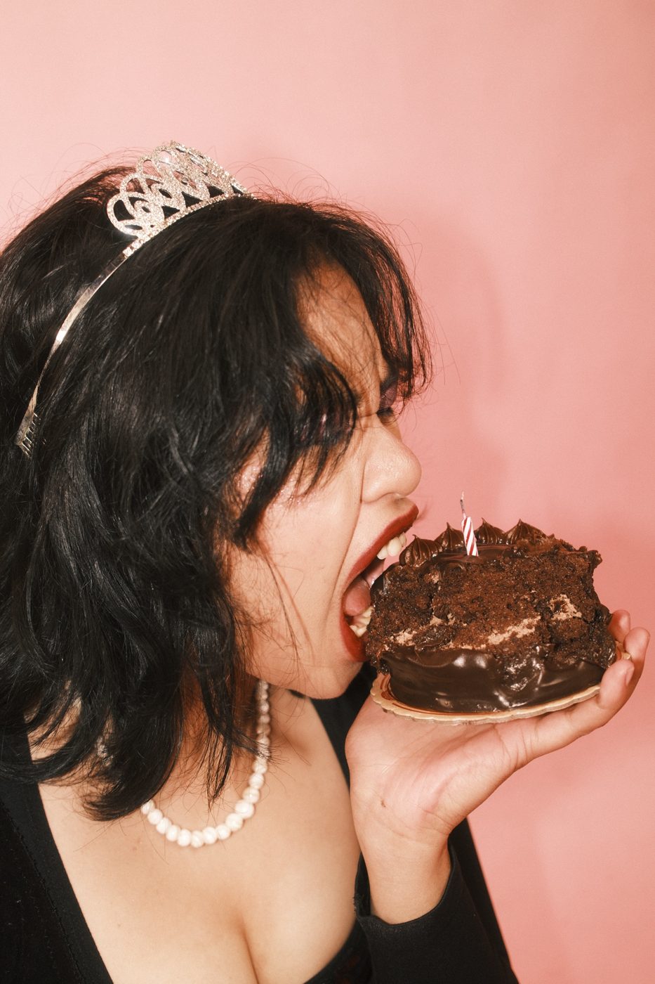 A woman in a tiara taking a huge bite of chocolate birthday cake against pink
