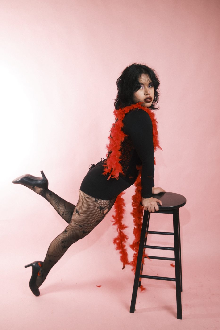 Leaning on a black stool in fishnets and a red feather boa, one heel kicked up