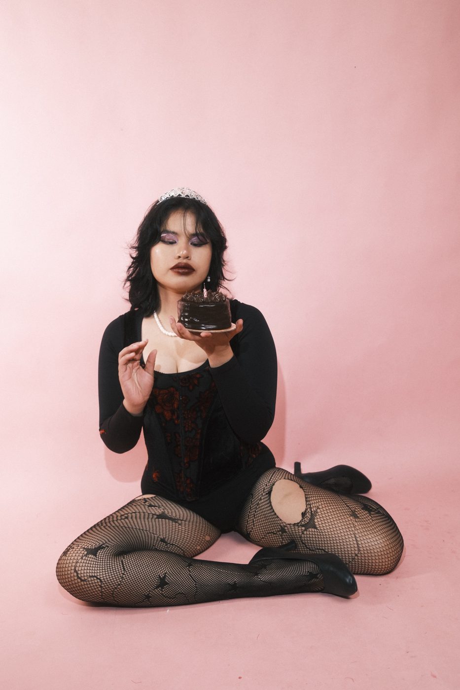 Seated on a pink floor in a tiara, studying a small chocolate cake