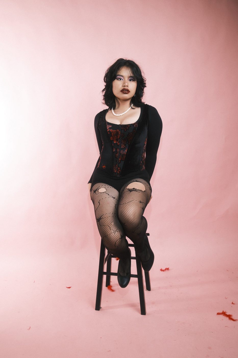 Perched on a stool in a velvet corset, pearls and torn fishnets