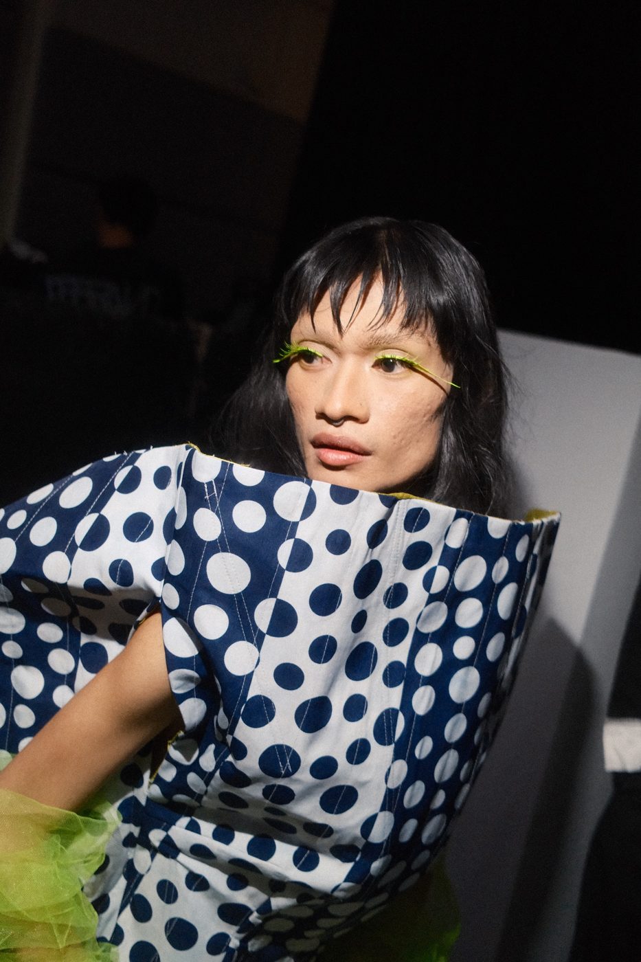 Model in a sculptural polka-dot dress with neon green lashes, half lit in the dark
