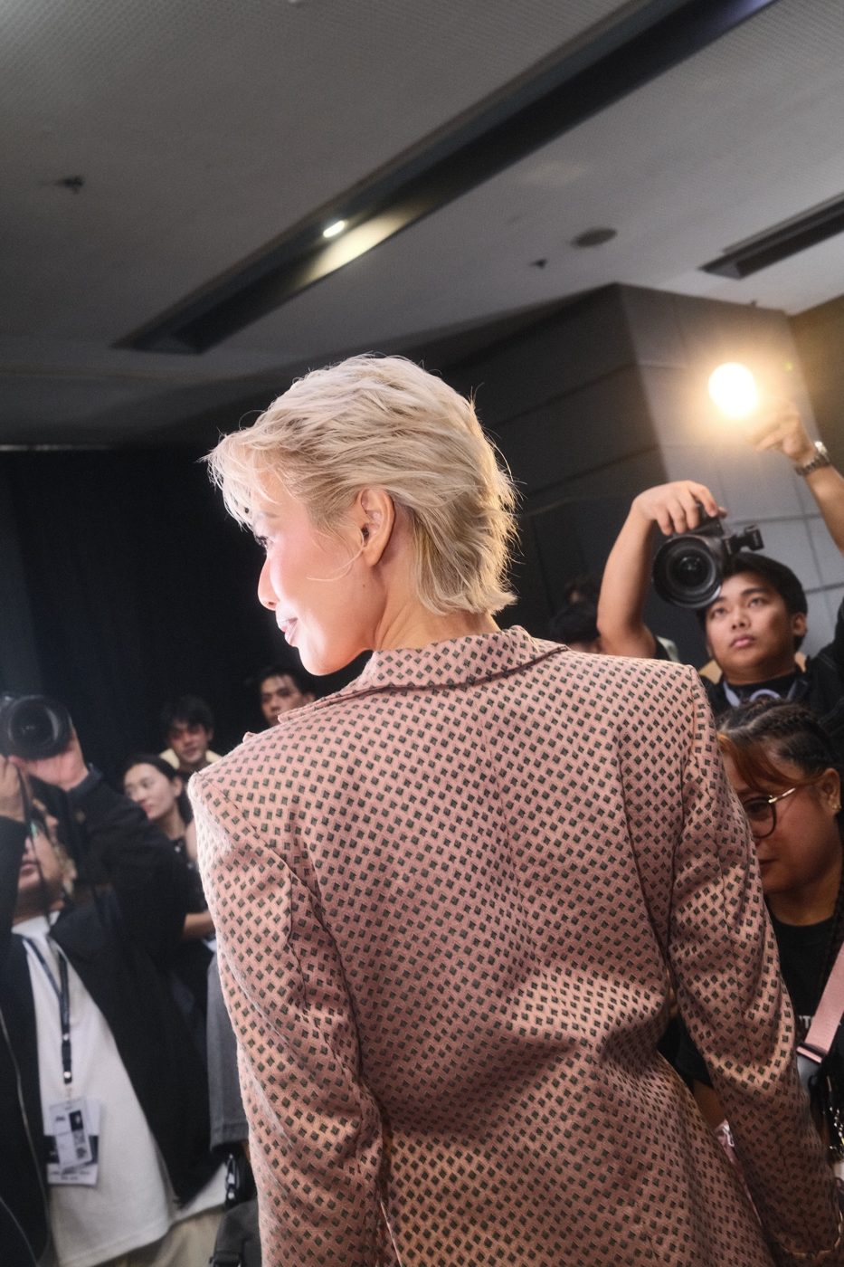 Blonde model seen from behind, facing a wall of photographers and a camera flash