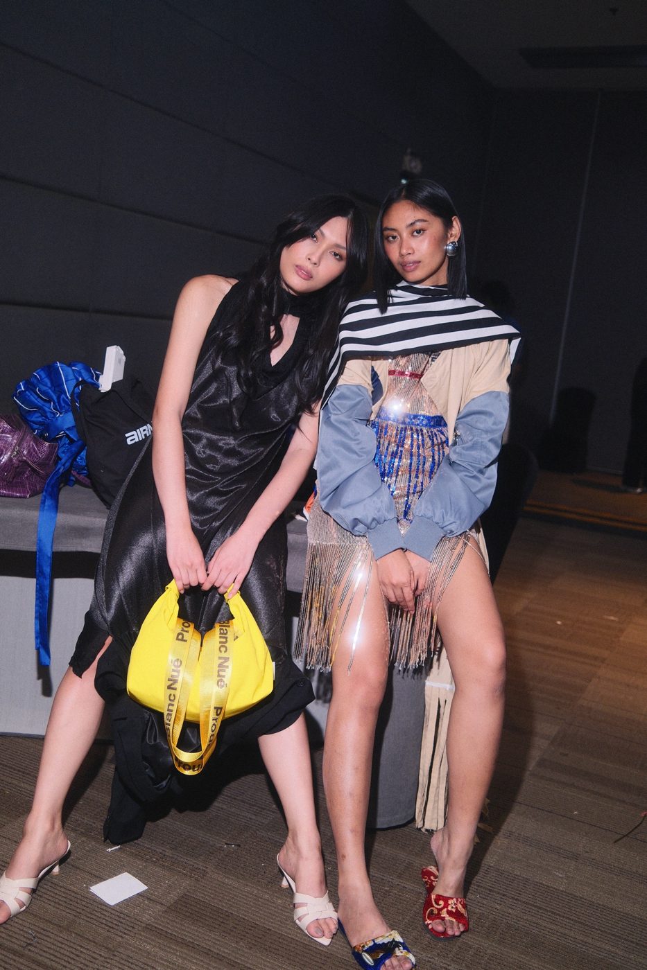 Two models leaning together backstage, one holding a bright yellow bag