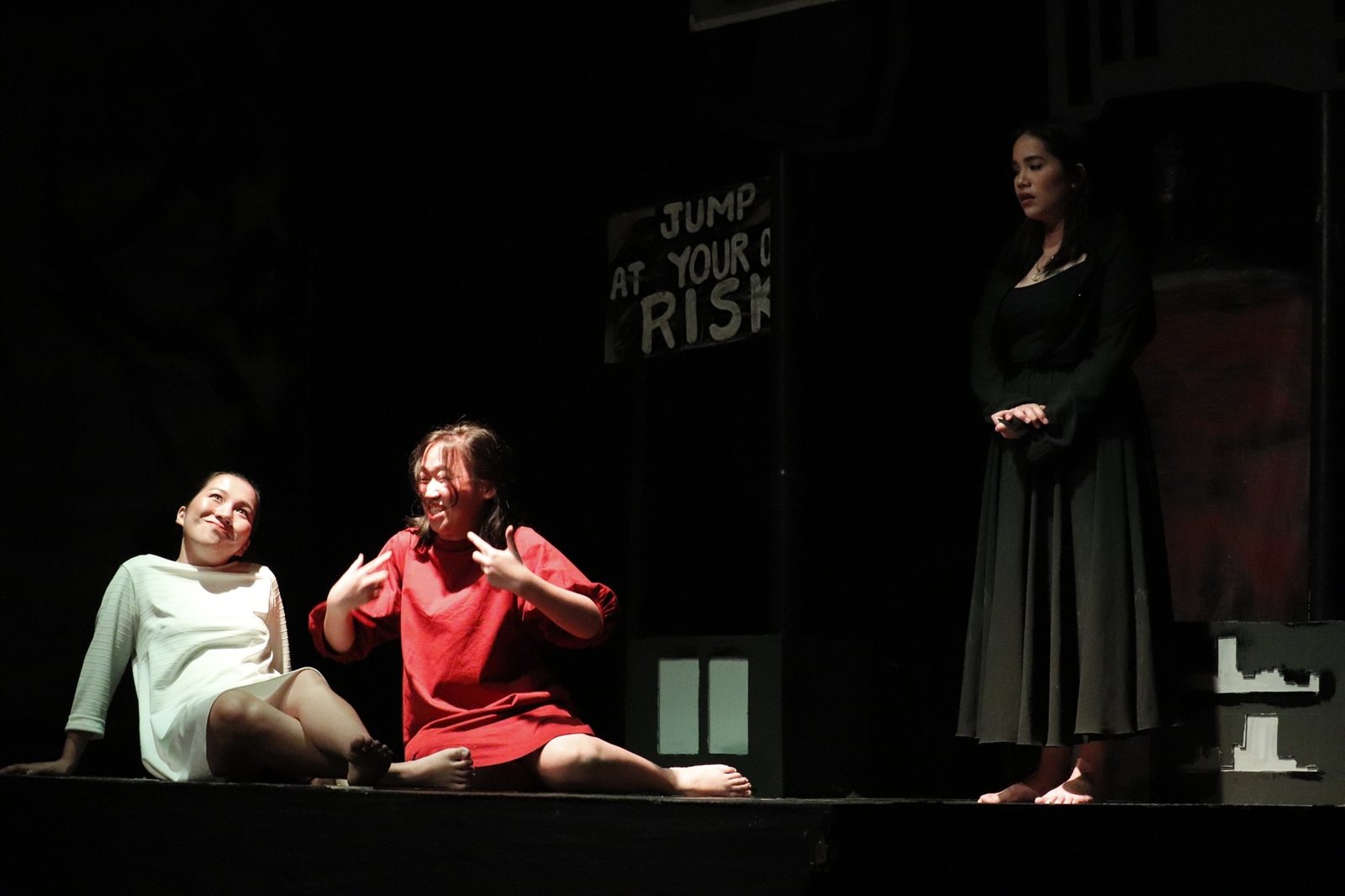 Three actors on a dark stage under a sign reading Jump at your own risk