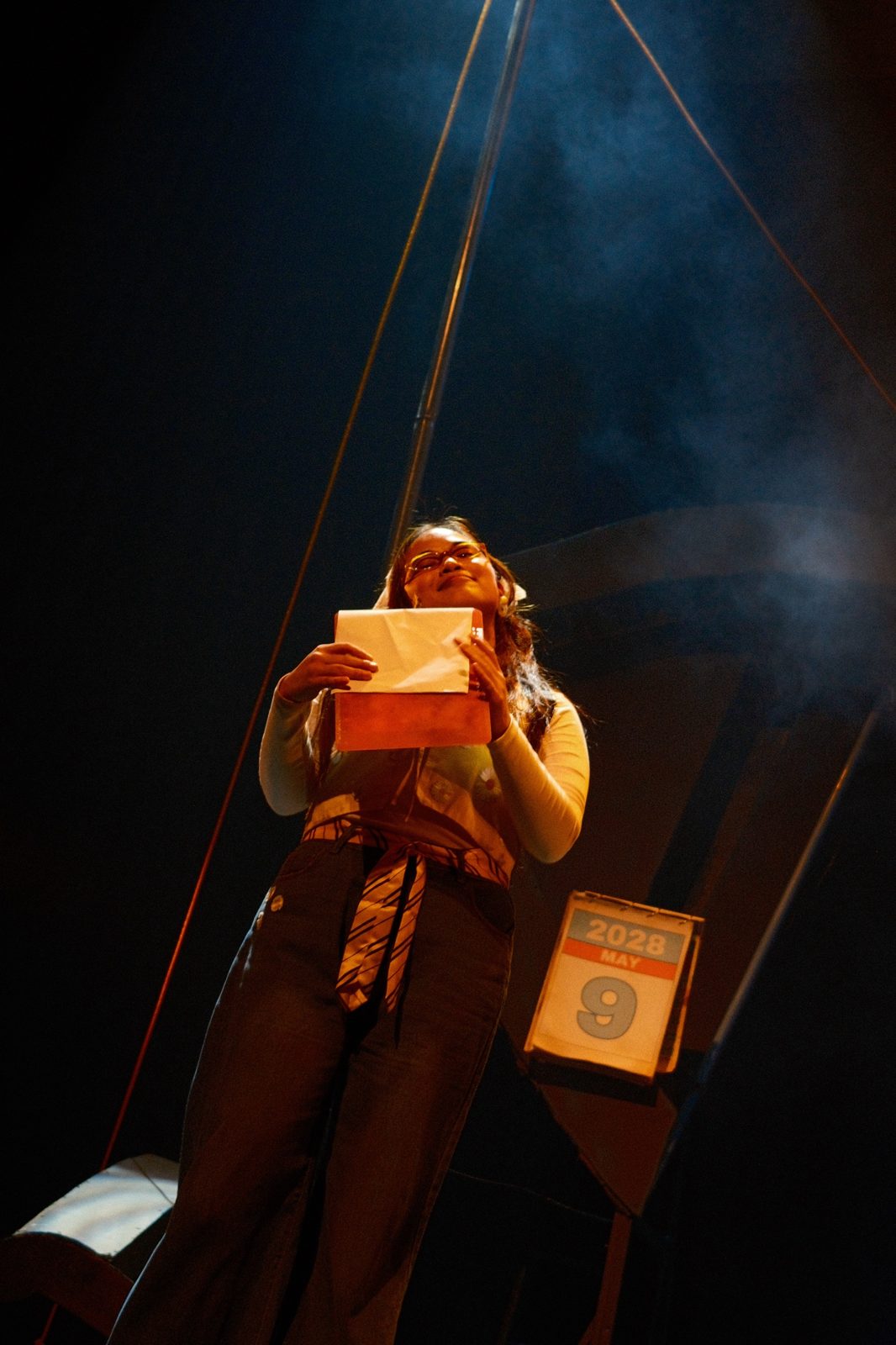 An actor in glasses lit from below, holding up a letter beside a calendar reading May 9, 2028