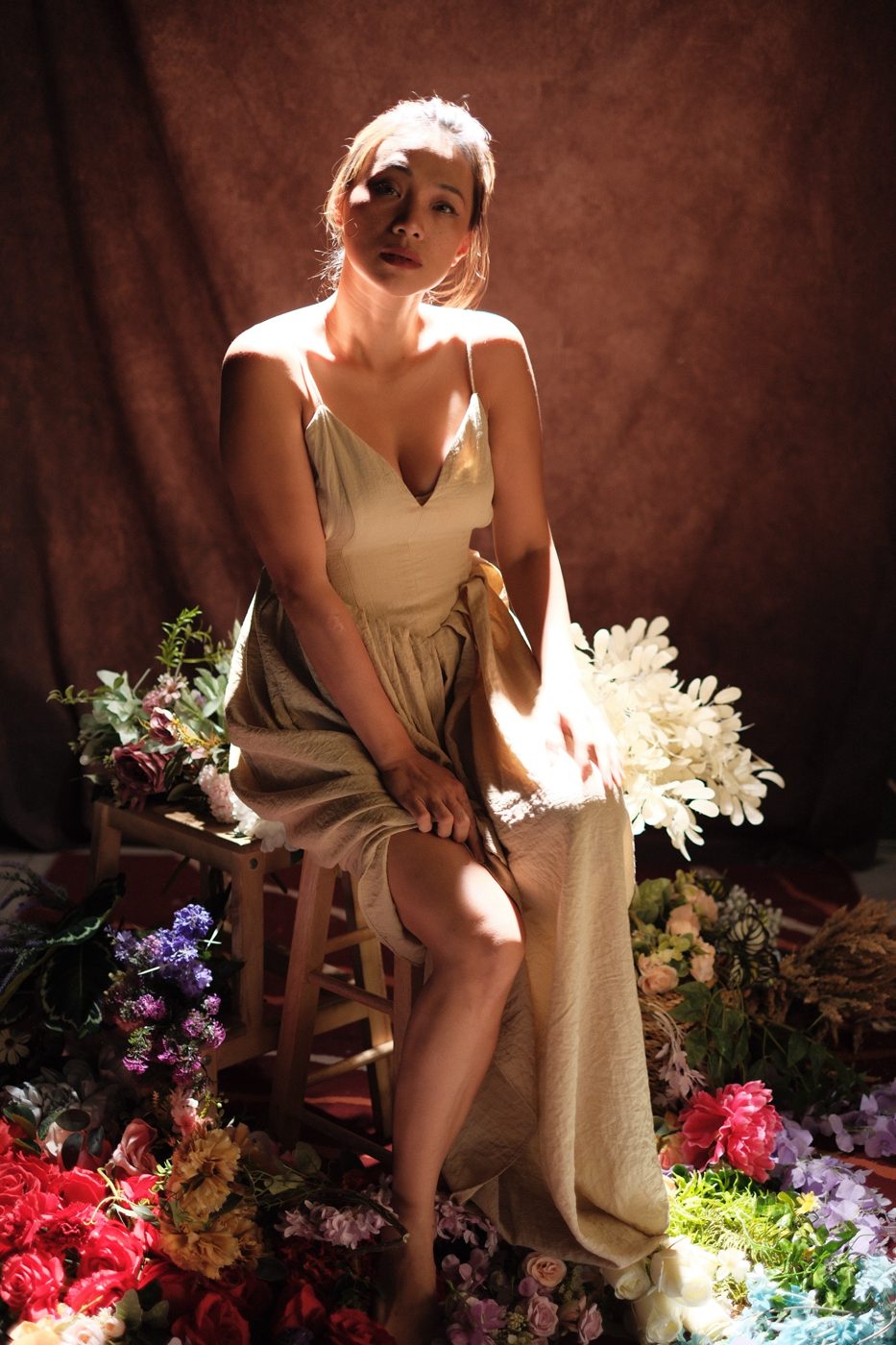 A woman in a linen dress seated on a stool, lit by a shaft of sun among flowers
